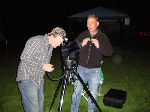 Two men set up a telescope outdoors at night, one adjusting the device and the other preparing.